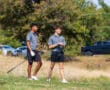 Two young golfers stand on the course, holding clubs, on a sunny day with trees in the background.