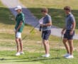 Three golfers in shorts and shirts on a putting green, holding putters, with a golf cart path in the background.