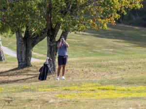 Golfer using rangefinder on a sunny course, surrounded by trees, with golf bag nearby.