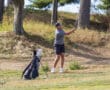 Golfer taking a swing on a sunny day, golf bag nearby on a grassy course, surrounded by trees.