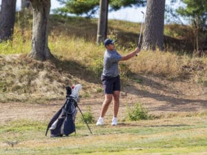 Golfer taking a swing on a sunny day, golf bag nearby on a grassy course, surrounded by trees.