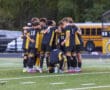 Soccer team huddling before a match on the field with Go Tigers bus in the background.
