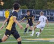 Youth soccer match in action, player in black and yellow jersey dribbles past opponent in white on a green field.