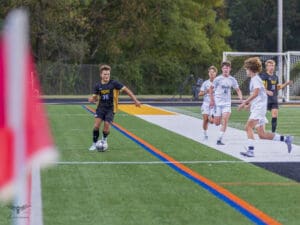Soccer player in black uniform dribbles the ball along the sideline, pursued by opponents in white uniforms.
