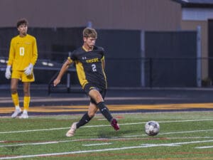 Soccer player in black uniform kicks ball during match, with goalie in yellow in the background.
