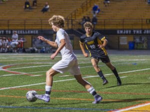 Soccer player in white dribbles the ball while a player in black chases on a brightly lit field.