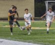 High school soccer match action, player in black dribbles ball while white team players chase, on a well-lit field.