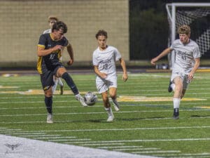 High school soccer match action, player in black dribbles ball while white team players chase, on a well-lit field.