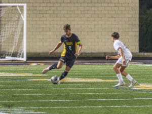 Two soccer players compete for the ball on a field during a night match, one wearing a black Tigers jersey.
