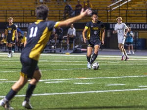 Soccer players in competitive match, focus on player dribbling ball on green field under stadium lights.