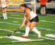Youth field hockey player in action, wearing white and black uniform, hitting a bright ball on artificial turf.