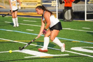 Youth field hockey player in action, wearing white and black uniform, hitting a bright ball on artificial turf.