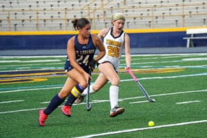 Two field hockey players compete for the ball on a green turf field.