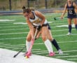 Field hockey player in action, wearing white and blue, preparing to hit yellow ball on a turf field.