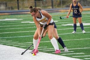 Field hockey player in action, wearing white and blue, preparing to hit yellow ball on a turf field.