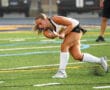 Field hockey player in action swinging stick on a turf field, wearing white socks and black shorts.