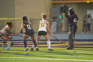 Field hockey match action with players aiming to score a goal, goalie ready to defend the net.