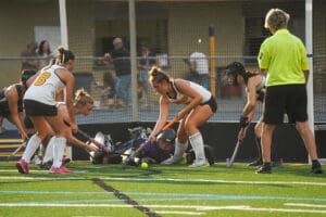 Field hockey players in action at the goal, with a referee observing closely during a competitive match.