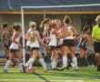 Girls' field hockey team celebrating a goal on the field with referee nearby.