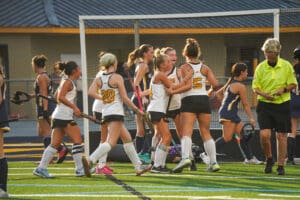 Girls' field hockey team celebrating a goal on the field with referee nearby.