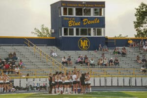 High school field hockey game at Mt. Lebanon Stadium, Blue Devils, with players huddled and spectators in stands.