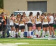 Girls' field hockey team huddles during a game, wearing matching uniforms with numbers, on a green turf field.