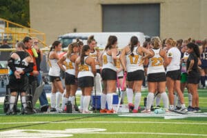 Girls' field hockey team huddles during a game, wearing matching uniforms with numbers, on a green turf field.