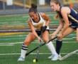 Two field hockey players in action on a green field. One wears a Tigers jersey, while the other wears blue.