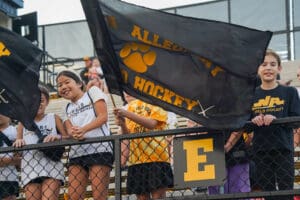Youth field hockey fans cheer with flags at a game in stadium stands.