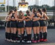 Field hockey team huddle before game at stadium with scoreboard in background.