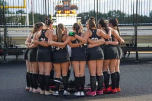 Field hockey team huddle before game at stadium with scoreboard in background.