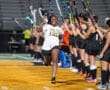 Smiling athlete runs through cheering teammates holding hockey sticks on a brightly lit field.