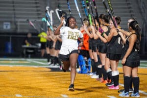 Smiling athlete runs through cheering teammates holding hockey sticks on a brightly lit field.