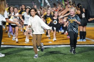 Children excitedly run through a group high-five line at a sports event on a turf field.