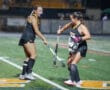 Two female field hockey players in a match at night on a green pitch, wearing black uniforms with sticks.