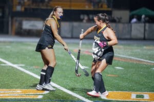 Two female field hockey players in a match at night on a green pitch, wearing black uniforms with sticks.