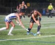 Two female field hockey players in action, competing for the ball on a bright, green turf field.