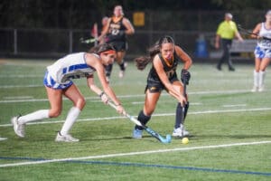 Two female field hockey players in action, competing for the ball on a bright, green turf field.