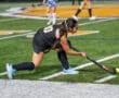 Field hockey player in black uniform preparing to hit ball during night game on artificial turf field.