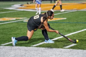 Field hockey player in black uniform preparing to hit ball during night game on artificial turf field.