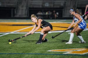 Two female field hockey players in action on a turf field during a competitive night match.