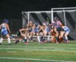 Girls' field hockey match at night, players from opposing teams competing for the ball near the goal.