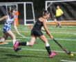 Two female field hockey players competing in a match, one dribbling the ball on a green turf under stadium lights.