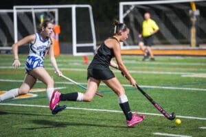 Two female field hockey players competing in a match, one dribbling the ball on a green turf under stadium lights.