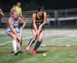 Two female field hockey players from opposing teams competing for the ball on a grass field at night.
