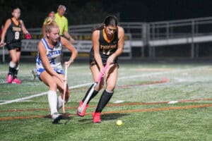 Two female field hockey players from opposing teams competing for the ball on a grass field at night.