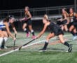 High school girls' field hockey match at night, players in motion on the turf, intense gameplay scene.
