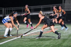 High school girls' field hockey match at night, players in motion on the turf, intense gameplay scene.