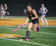 Field hockey player in action at night, dribbling a ball with a pink stick on a green turf field.
