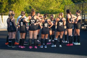 Field hockey team huddles during practice, wearing black uniforms with numbers, under a sunny sky.